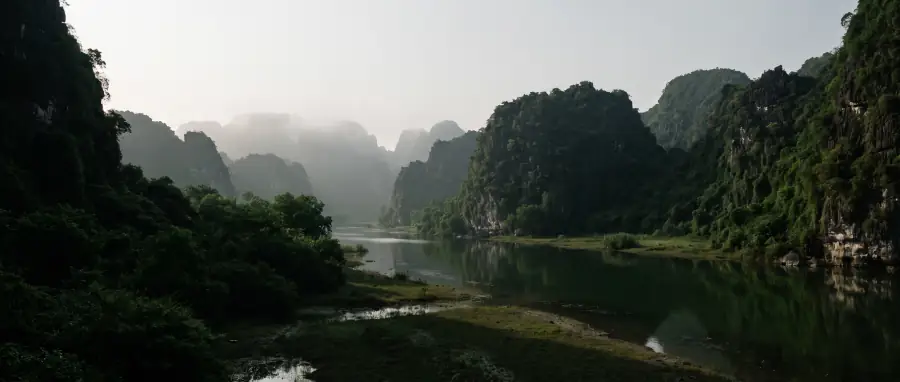 Still river water between limestone cliffs at Ninh Binh in early morning, mist on the far cliffs and dark green vegetation reflected in the water.