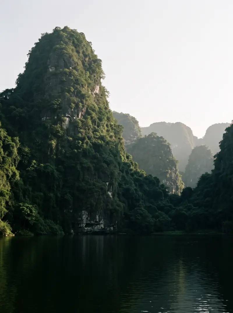 Limestone karst towers rising from still water at Ninh Binh, late afternoon light raking from the left and throwing long shadows across the water.