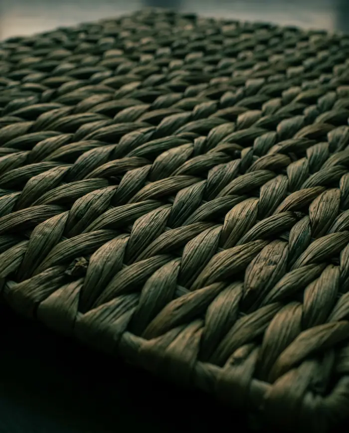 A woven panel of dried water hyacinth filling the frame, low side light raking across the weave so each strand throws its own shadow.